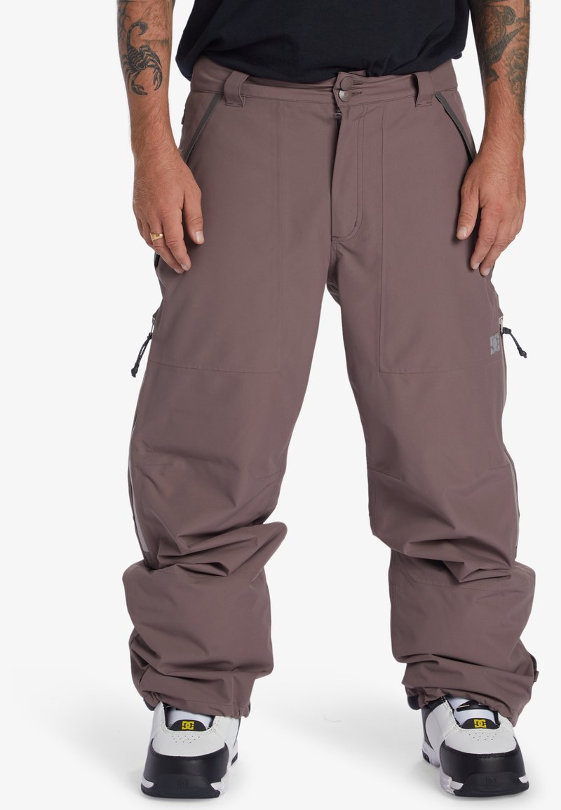 Pantalons neige légers, mauves, avec une coupe ample, plusieurs poches zippées et des poignets ajustables, associés à des baskets noir et blanc.