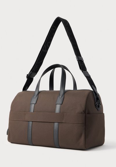 Καφέ ορθογώνια τσάντα duffel με μαύρες δερμάτινες λαβές και ρυθμιζόμενο μαύρο ιμάντα ώμου, εμφανιζόμενη σε λευκό φόντο.