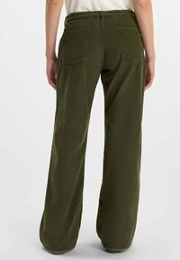 Vista trasera de una persona que lleva pantalones de pana anchos color verde oliva con dos bolsillos traseros y una blusa blanca.