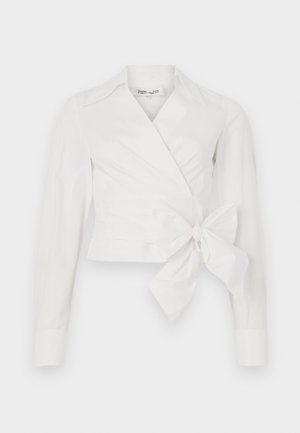 Blouse blanche à manches longues, col et détail de nœud à l'avant. Fabriquée en tissu lisse avec un design légèrement structuré.