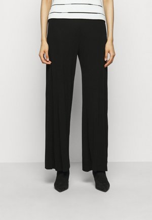 Frau trägt schwarze, weit geschnittene Hosen, schwarze spitze Stiefel und ein weißes ärmelloses Top mit dünnen horizontalen schwarzen Streifen.