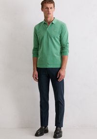 Mann in einem grünen Langarm-Polohemd, blauen Hosen und schwarzen Slippern, der vor einer schlichten weißen Wand steht.