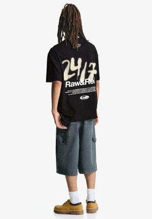 Black short-sleeve t-shirt with white graphic "24/7 Raw&Rel" on back, paired with gray denim shorts and yellow shoes. Casual design.