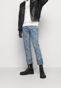 Blå denimjeans med ett mönster av loggor över hela, tillsammans med en svart läderbomberjacka och svarta kängor.