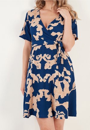 Femme portant une robe portefeuille bleue avec des motifs abstraits beiges et des manches courtes, touchant ses cheveux blonds ondulés.