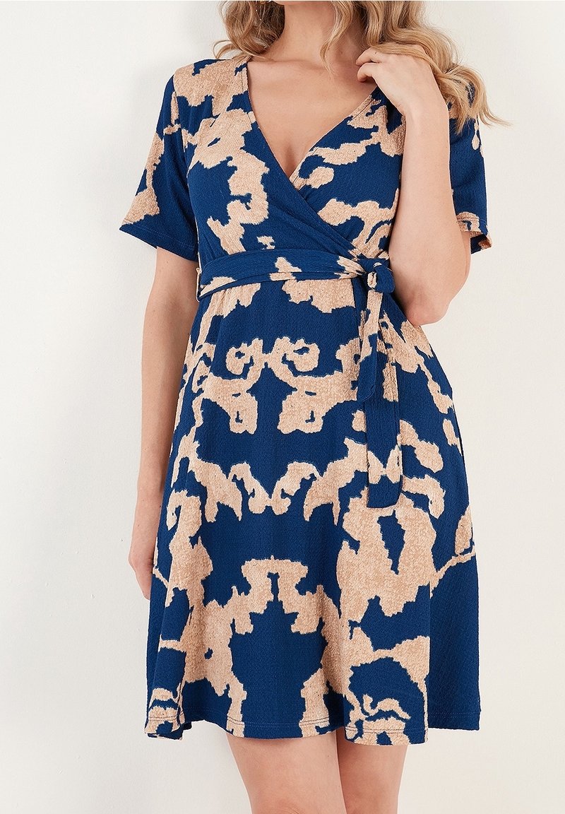 Femme portant une robe portefeuille bleue avec des motifs abstraits beiges et des manches courtes, touchant ses cheveux blonds ondulés.