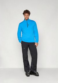 Pulôver azul de manga comprida com gola de meio fecho, combinado com calças pretas com bolsos com fecho e calçado preto, em pé contra um fundo claro.