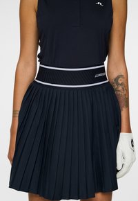 Mekko golfia sinivalkoisilla halkioilla, jossa on leveä vyötärö, ja jossa näkyy vinoraitoja. Valkoinen hansikas kädessä. Rakenteellinen kangas.