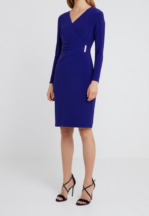 Vrouw die een knielange, lange mouwen, koningsblauwe wikkeljurk draagt met een zilveren gesp en zwarte sandalen met hoge hakken, staand op een witte vloer.