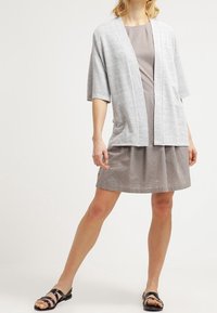 Cardigan in stile kimono grigio chiaro sopra un vestito a pois marrone. Il cardigan presenta maniche larghe e una vestibilità aperta, abbinato a sandali neri.