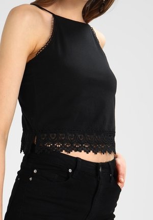 Frau trägt ein schwarzes ärmelloses Crop-Top mit Spitzensaum und schwarze hoch taillierte Hose, Hand in der Tasche.