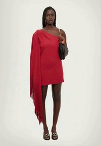 Rdeča obleka z enim ramenom in detajlom fringa, ki se spušča po eni strani. Manekenka drži črno torbico in nosi odprte čevlje s srčastimi akcenti.