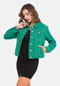 Grüne Jeansjacke mit weißen Nähten, zwei Brusttaschen und silbernen Knöpfen. Kombiniert mit einem schwarzen Kleid darunter.