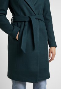 Mørk teal uldfrakke med bælte, båret lukket, med den ene hånd i lommen og synlige lyseblå jeans under kanten.