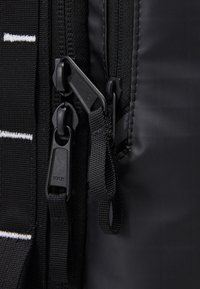 Deux fermetures éclair noires YKK avec languettes en tissu sur un sac noir, coutures visibles et tissu texturé à côté d'un matériau lisse.