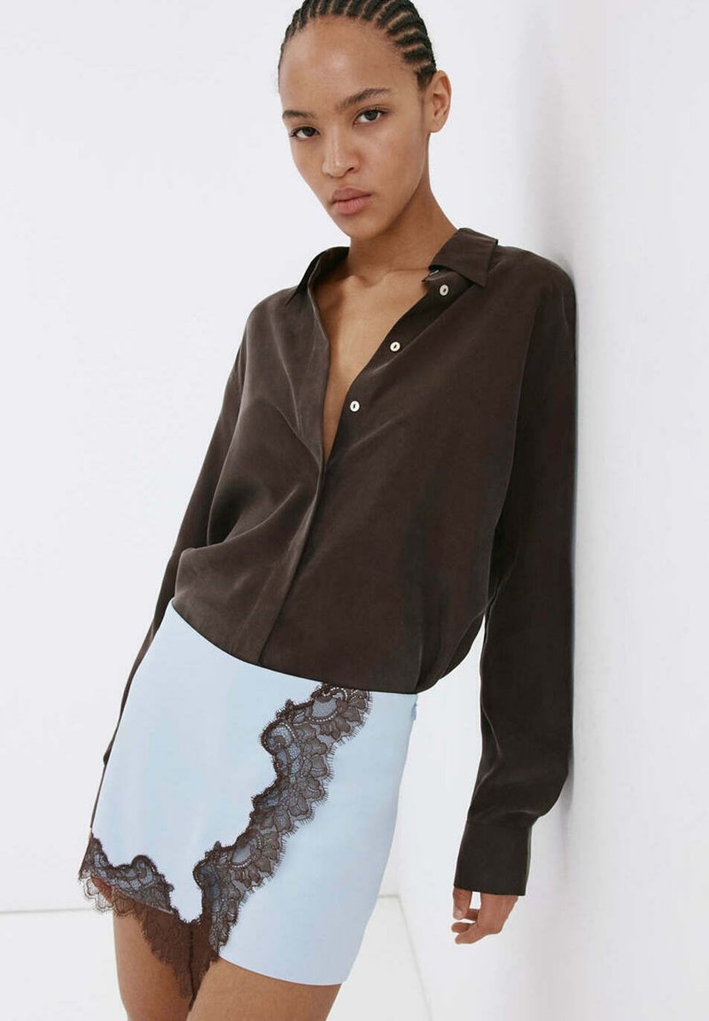 Femme aux cheveux tressés portant une chemise ample marron foncé boutonnée et une mini-jupe bleu clair avec un bord en dentelle noire, appuyée contre un mur blanc.