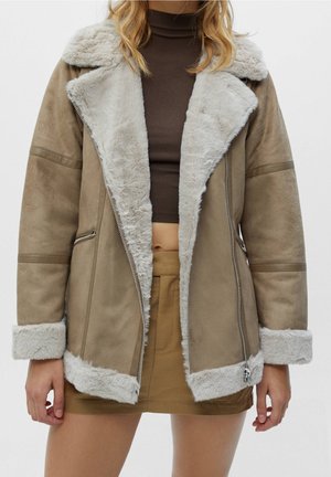 Nainen pukeutuneena vaaleanruskeaan karvahameiseen takkiin, jossa on valkoinen turkisreunus, päällään ruskea lyhyt toppi ja vaaleanruskea minihame, seisomassa yksivärisen taustan edessä.