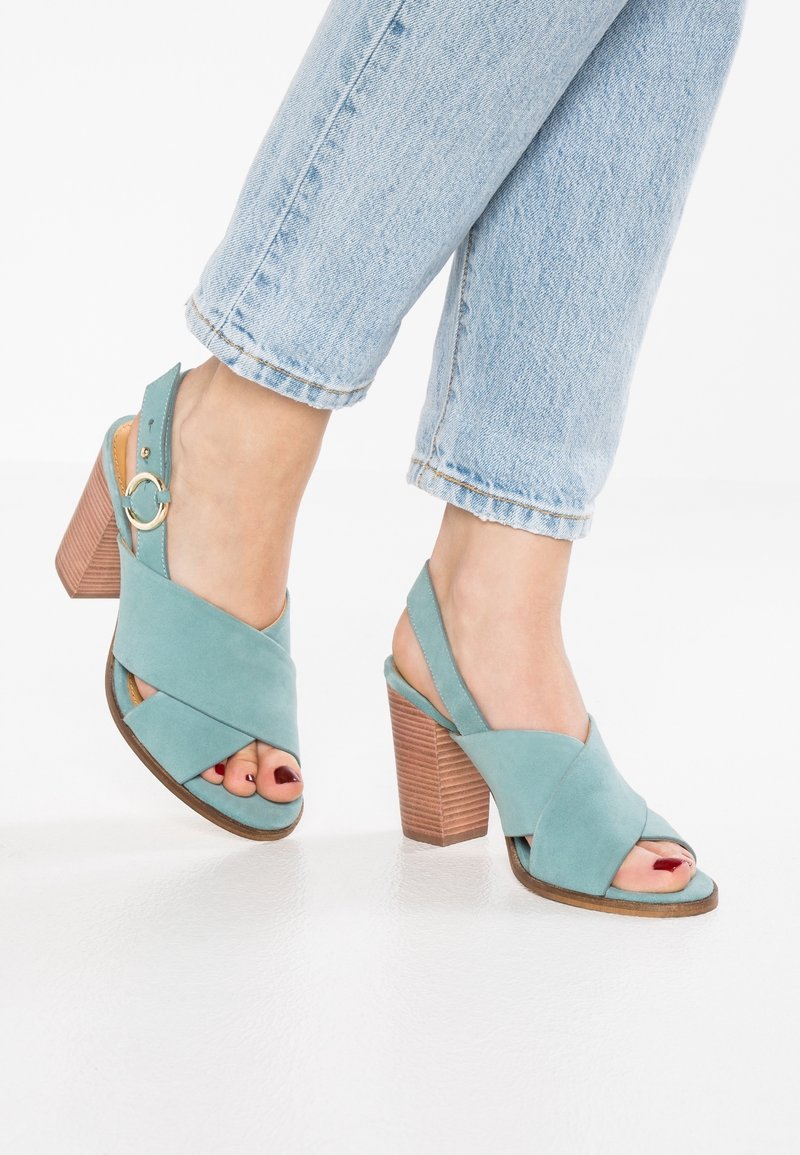 Sandales à talons en daim bleu sarcelle avec des brides croisées, bout ouvert et talon bloc en bois. La bride réglable est ornée d'un accent métallique circulaire.
