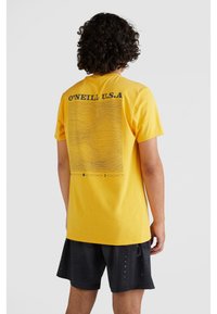 Camiseta de algodón amarilla con el texto negro "O'NEILL U.S.A." y un diseño de línea ondulada en la parte posterior, combinada con pantalones cortos negros con una textura estampada.