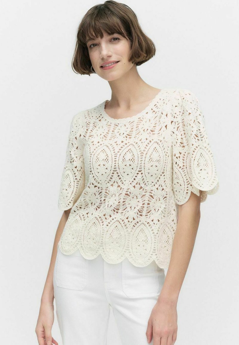 Lace topp i krämfärg med vågig nederkant, med blommönster och korta ärmar. Kombinerad med vita byxor.