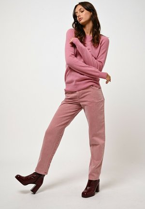 Maglione rosa realizzato in morbido tessuto, abbinato a jeans rosa chiaro. Il modello indossa stivaletti con tacchi in bordeaux lucido, in una posa dinamica.