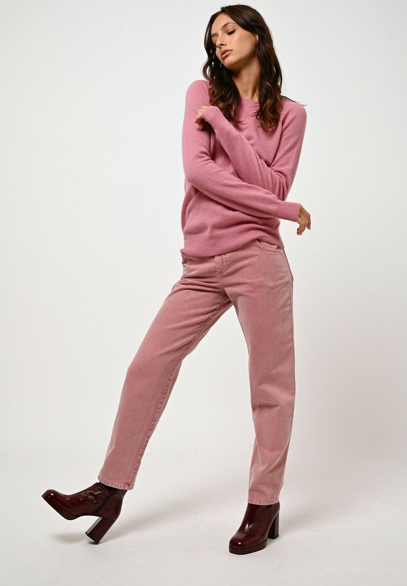 Maglione rosa realizzato in morbido tessuto, abbinato a jeans rosa chiaro. Il modello indossa stivaletti con tacchi in bordeaux lucido, in una posa dinamica.