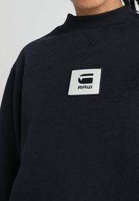 Marina sweatshirts i bomullsmaterial, med rund halsringning och en vit rektangulär patch med "RAW"-logotypen på framsidan.