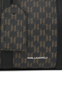 Melnas ādas soma ar atkārtotu brūno logotipa rakstu. Iekļauj priekšējo kabatu un sudraba zīmola nosaukuma plāksni ar struktūru.