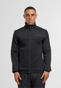 Černá zipová bunda vyrobená z měkké tkaniny, s vysokým límcem, texturovanými panely a decentním logem na levém horním rohu.