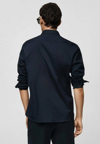 Camicetta blu navy con maniche arrotolate, realizzata in un tessuto morbido; presenta un colletto classico e un orlo curvo nella parte posteriore.