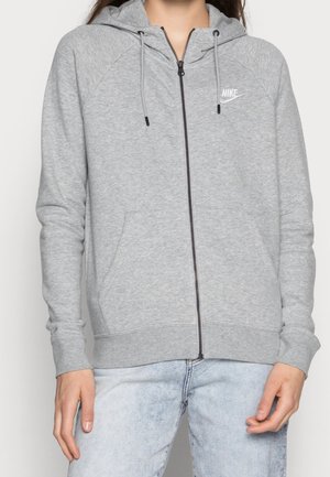 Personne portant un sweat à capuche zippé Nike gris clair avec cordons et poches avant, assorti à un jean en denim bleu clair.