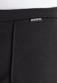 Gros plan sur un tissu noir texturé avec une petite étiquette noire affichant le texte blanc "HUGO" près du bord supérieur.