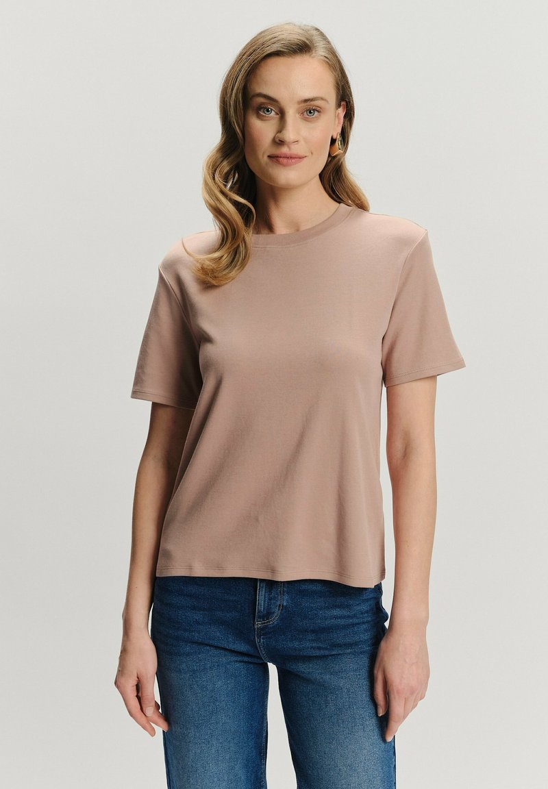 Kobieta z falującymi blond włosami, nosząca złote kolczyki w kształcie kół, jasno brązową koszulkę z krótkim rękawem i niebieskie dżinsy, stojąca na jednolitym tle.