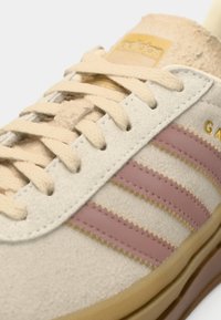 Baskets en suède beige avec trois bandes rose clair, lacets crème et semelle beige. Le branding doré sur la languette complète le design.