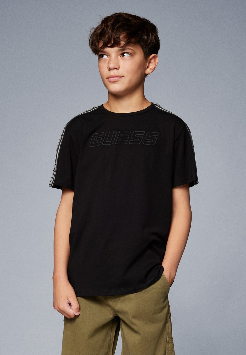 Svart bomull T-skjorte med et hevet "GUESS"-logo på forsiden, komplementert av stripete detaljer på ermmene. Løs passform.