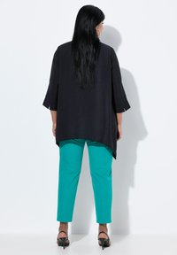 Tunic nera a maniche lunghe con orlo curvo, abbinata a pantaloni turchesi brillanti; il modello indossa tacchi aperti neri e un colletto aderente.