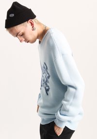 Maglione azzurro chiaro lavorato a maglia con vestibilità ampia, caratterizzato da una grafica in blu scuro. Indossato con un berretto nero e pantaloni neri.