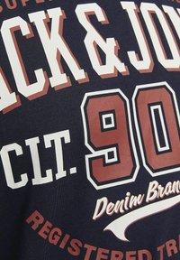 Mørkeblå T-shirt med store hvide og maroon tekst. Har "JACK & JONES," år "90," og "Denim Brand" i varierende skriftstørrelser og stilarter.