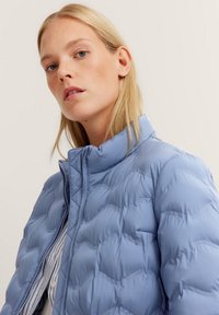 Hellblau gesteppte Puffjacke mit hohem Kragen, ausgestattet mit einem Frontreißverschluss und einem strukturierten, gepolsterten Design. Horizontale Nähte sind sichtbar.