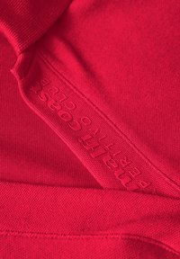 Červené sportovní tričko s hladkou texturou. Obsahuje vyražené logo podél švu a má přiléhavý design vhodný pro aktivní použití.