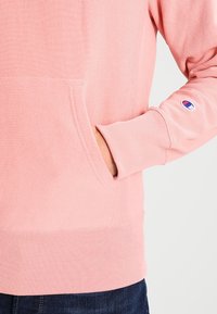 Lichtroze hoodie met een kangoeroezak, geribbelde manchetten en zoom. Voorzien van een logopatch op de linkermouw. Gemaakt van zachte stof.