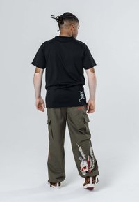 T-shirt in cotone nero con una vestibilità rilassata, abbinata a pantaloni cargo oliva con dettagli ricamati di teschio e ali.