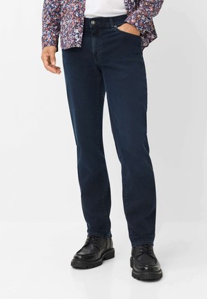 Mand iført mørkeblå jeans med lige ben, sorte snørebåndsstøvler, en hvid skjorte og en langærmet blomstret skjorte med den ene hånd i lommen.