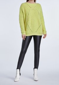 Pull jaune en maille avec des détails de points ajourés, associé à des leggings noirs et des bottines blanches. Le pull présente une coupe ample et des poignets côtelés.