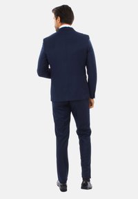 Abito blu navy con vestibilità adattata, dotato di uno spacco singolo, pantaloni a gamba dritta e scarpe nere lucide, presentato da dietro.