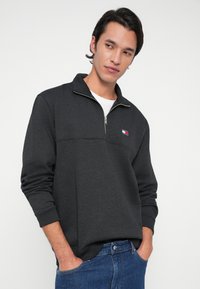 Νεαρός άνδρας με κοντά σκούρα μαλλιά, φορώντας σκούρο γκρι φούτερ με κουκούλα και μπλε τζην, με τα χέρια στις τσέπες, κοιτάζοντας αριστερά.