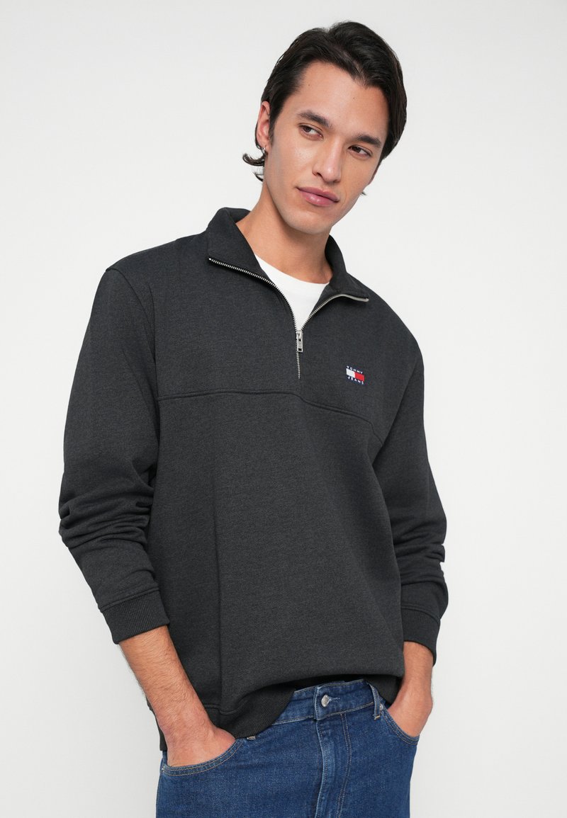 Junger Mann mit kurzen dunklen Haaren, der einen dunkelgrauen Quarter-Zip-Sweatshirt und blaue Jeans trägt, mit den Händen in den Taschen und nach links schaut.