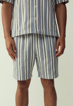 Mand iført blå og hvide lodrette stribede skjorte med knapper og matchende shorts, stående med armene afslappet ved siderne.