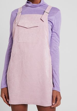 Woman wearing a light purple turtleneck under a pale pink corduroy pinafore dress with front flap pocket and adjustable straps.