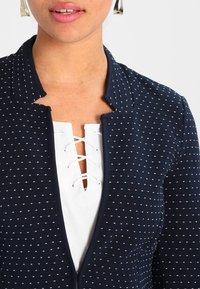 Giacca blu navy con texture a pois, colletto aperto e dettagli in pizzo bianco sul petto. Tessuto morbido con un design aderente.